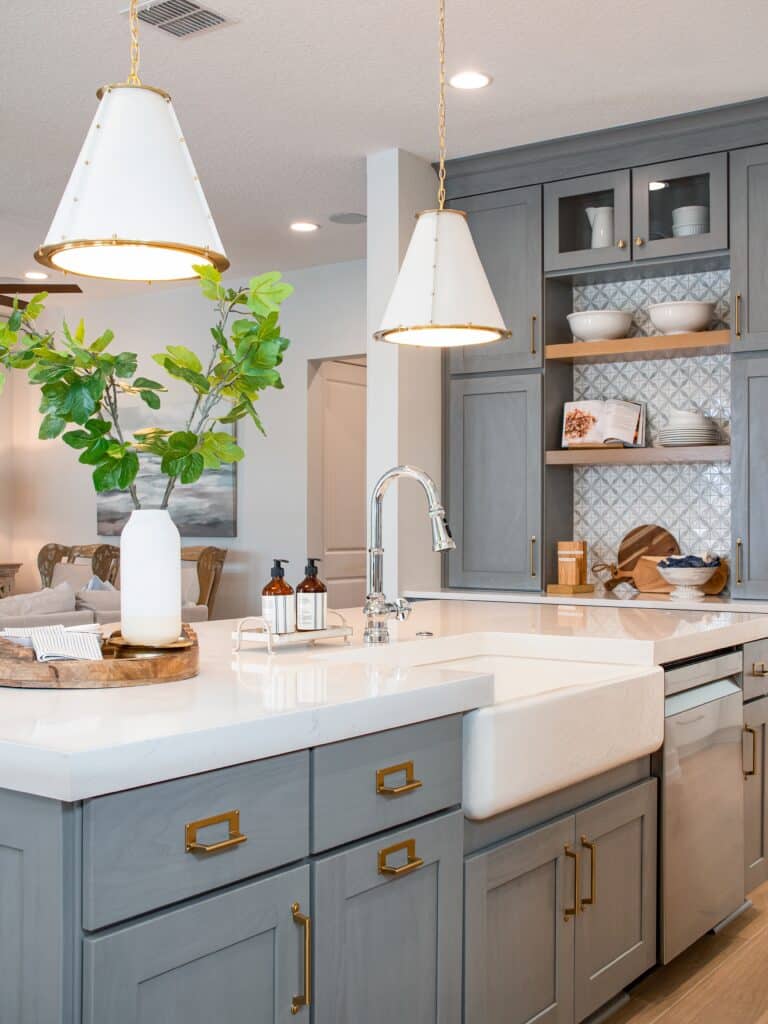 A bright kitchen with gray cabinetry, brass hardware, a white farmhouse sink, a quartz island countertop, cone pendant lights, and a patterned tile backsplash